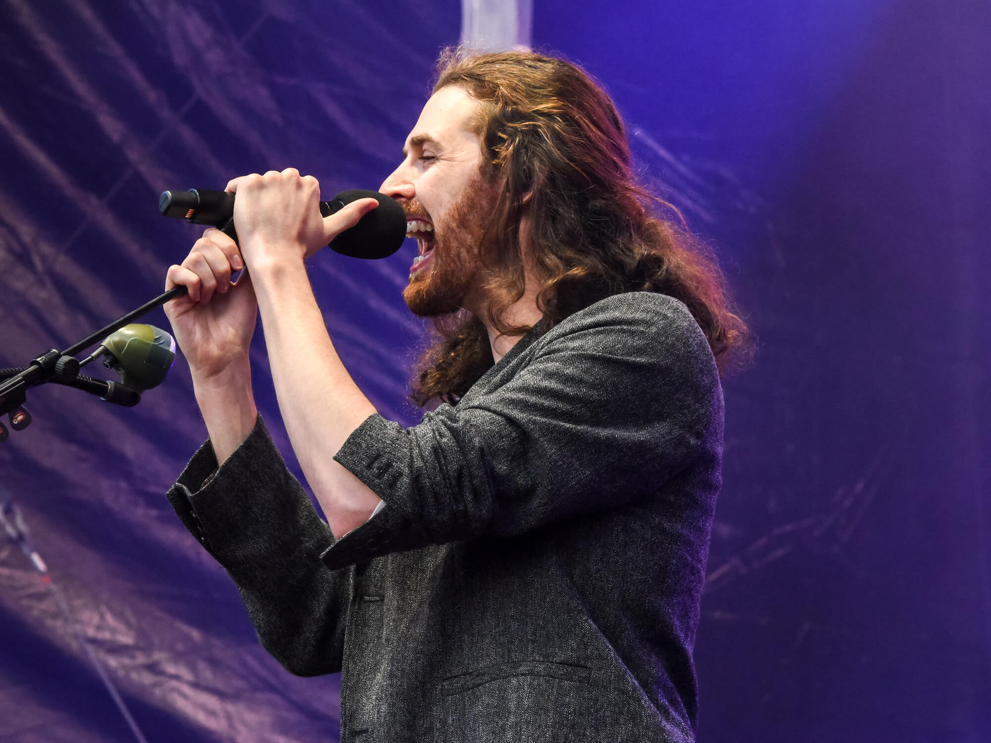 Hozier on stage, he is gripping on to a microphone on a stand and singing