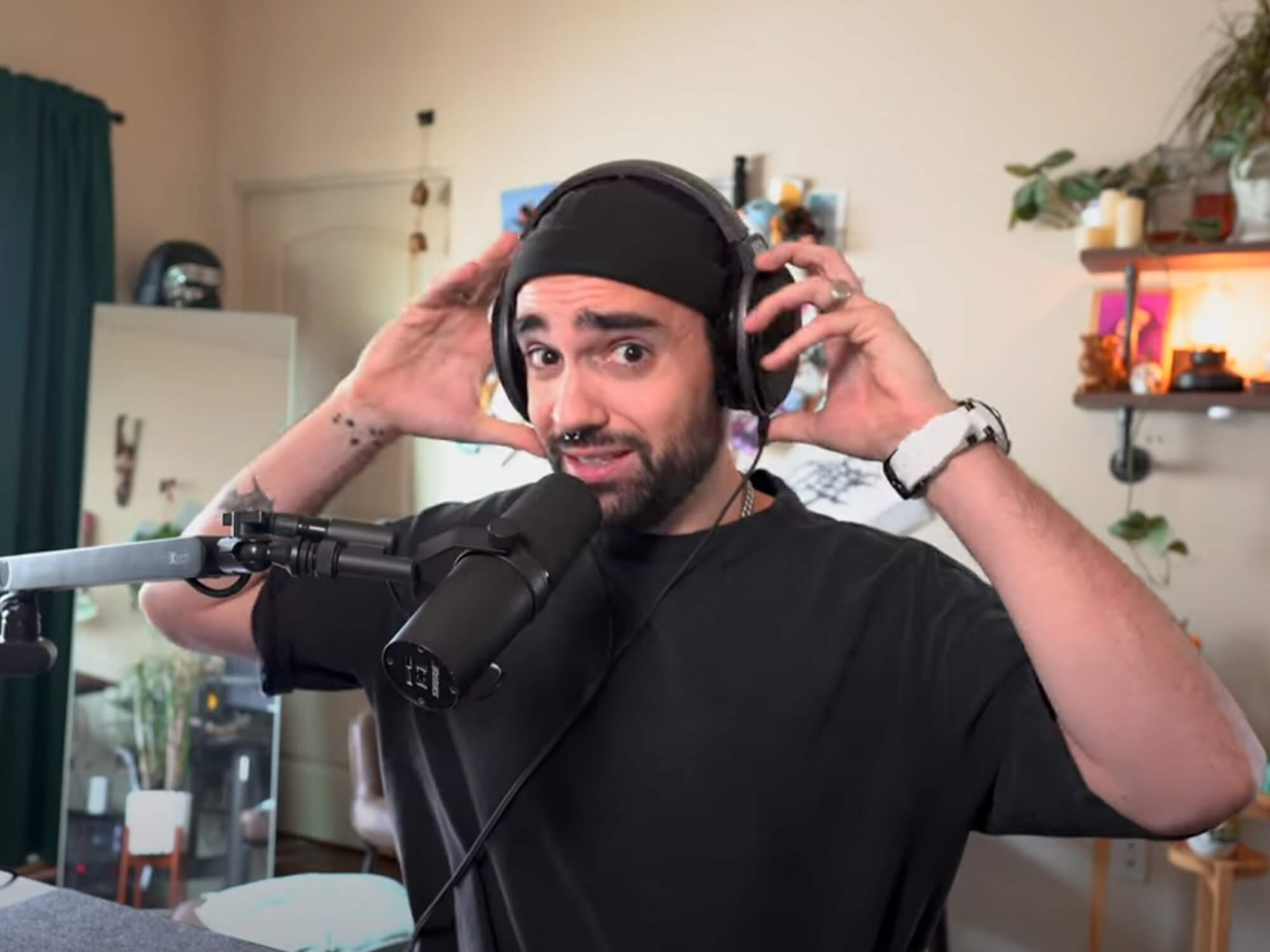 TAETRO in his YouTube video. He is placing headphones on his head and looks skeptical. He's wearing a black t-shirt and black beanie hat.