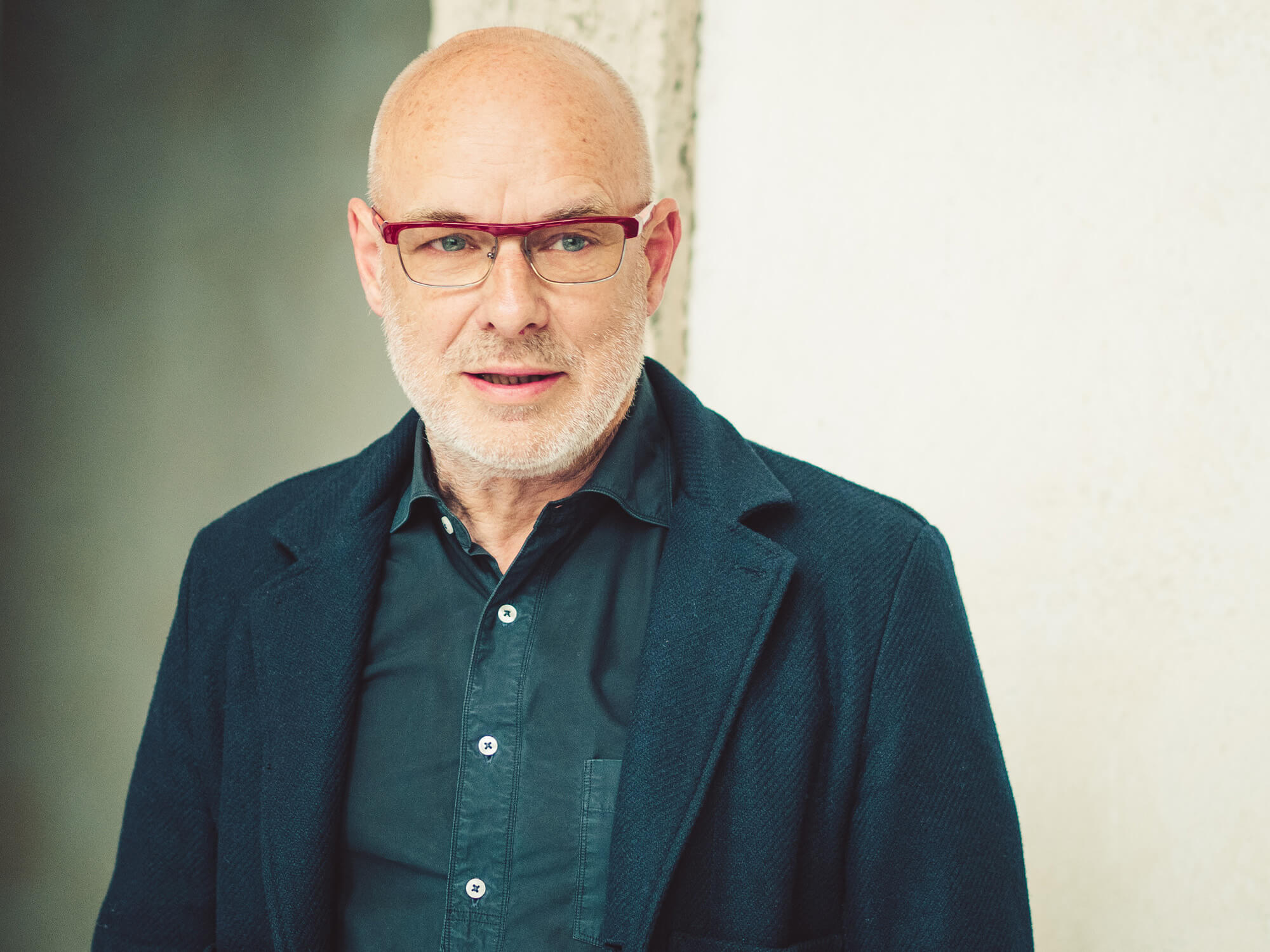 Brian Eno looking off to the right, with his mouth slightly open as if he is speaking. He wears red-framed glasses and a navy shirt and jacket.