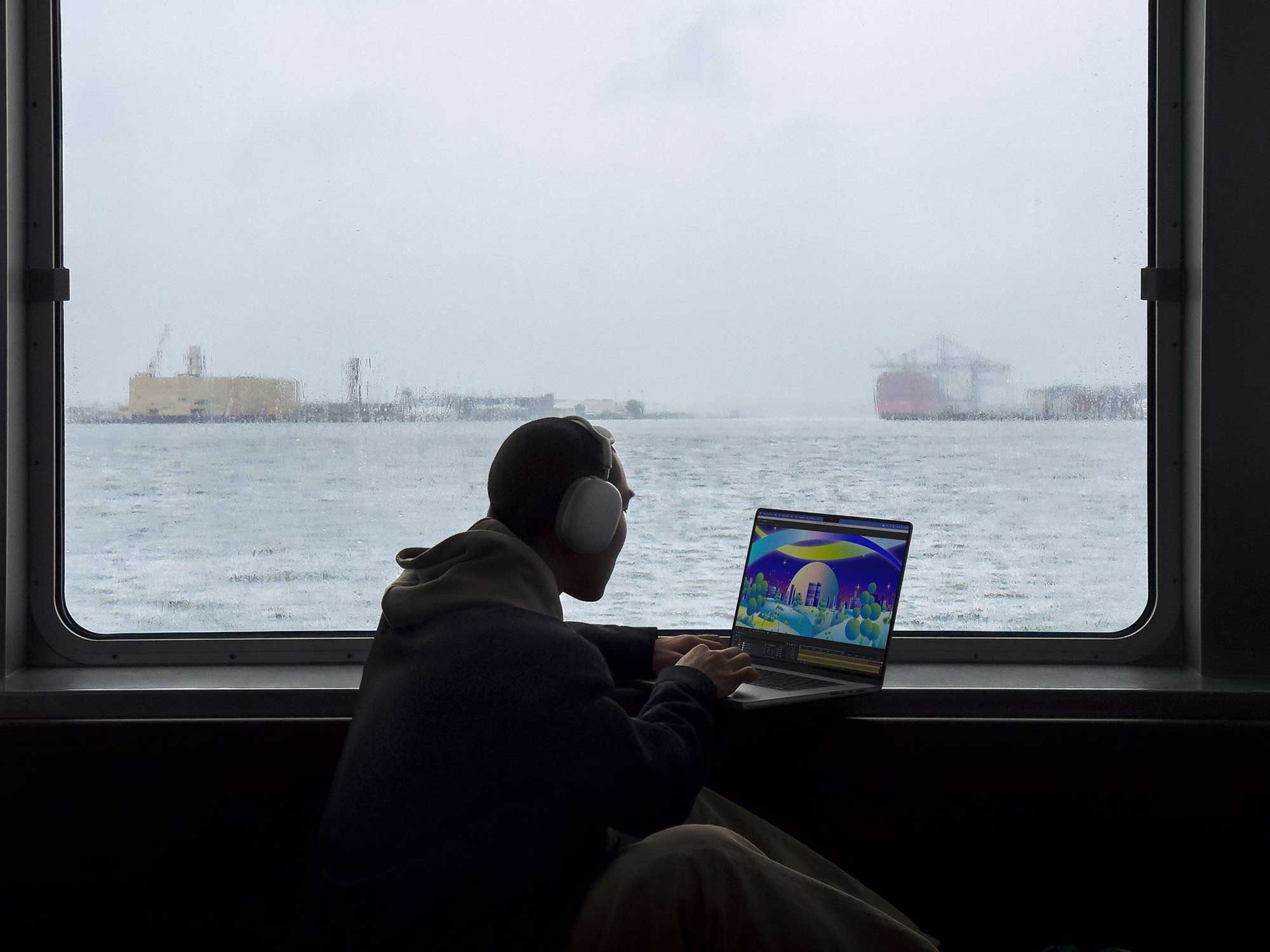 A man uses a MacBook Pro on a boat next to a window on a rainy day