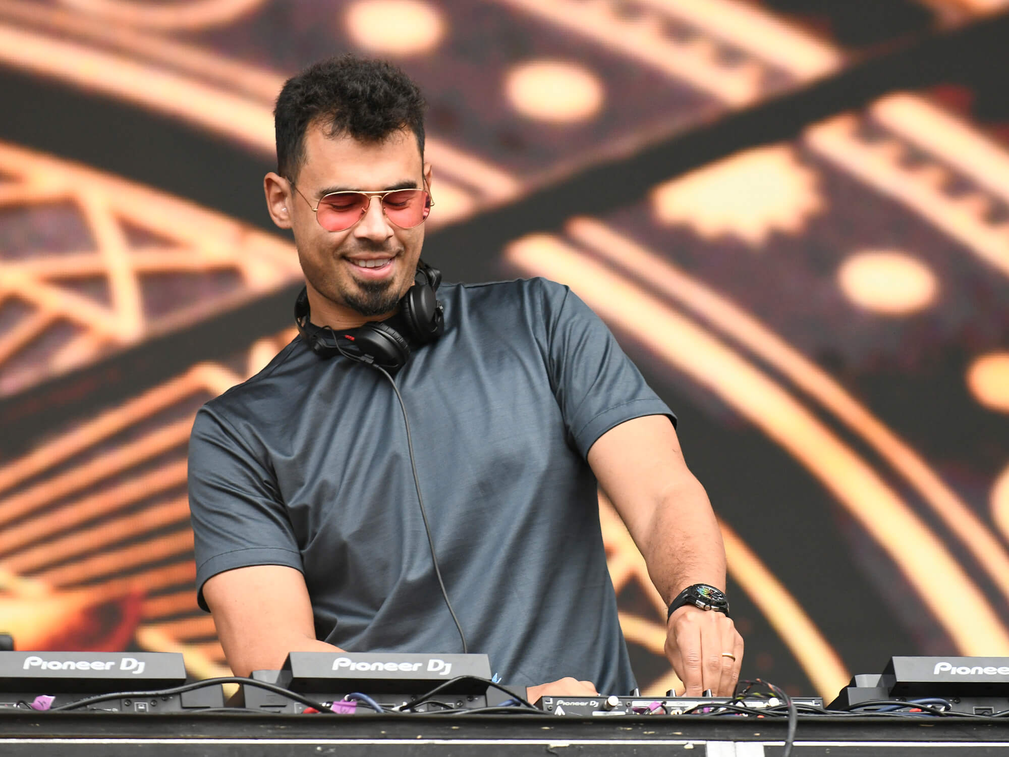 Afrojack behind the decks on stage in 2023. He wears headphones around his neck and has red-coloured sunglasses on. He's smiling and looking down.
