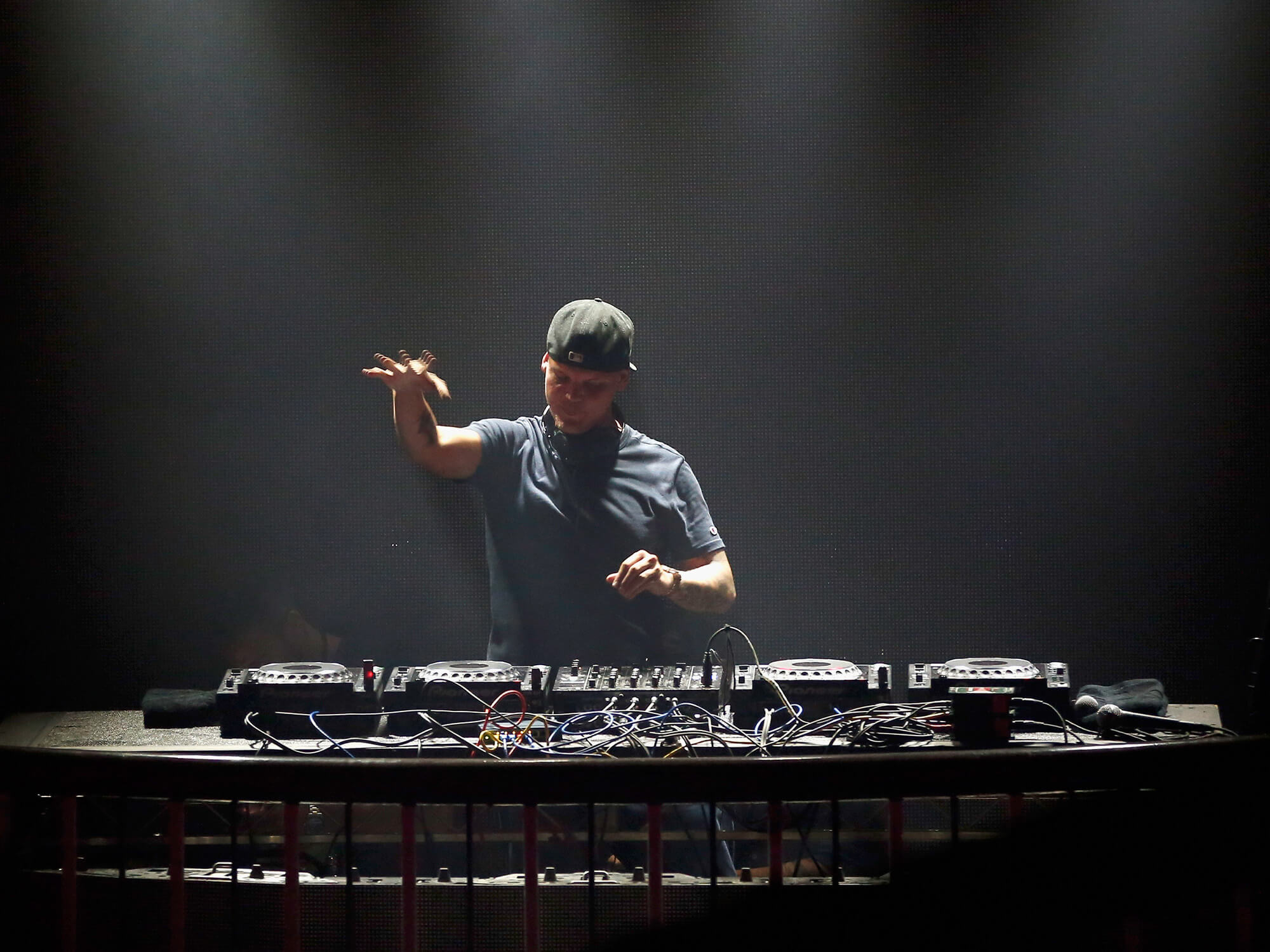 Avicii on stage in 2016. He is wearing a t-shirt and cap and has one hand hovering above the deck. White lighting shines down on him.