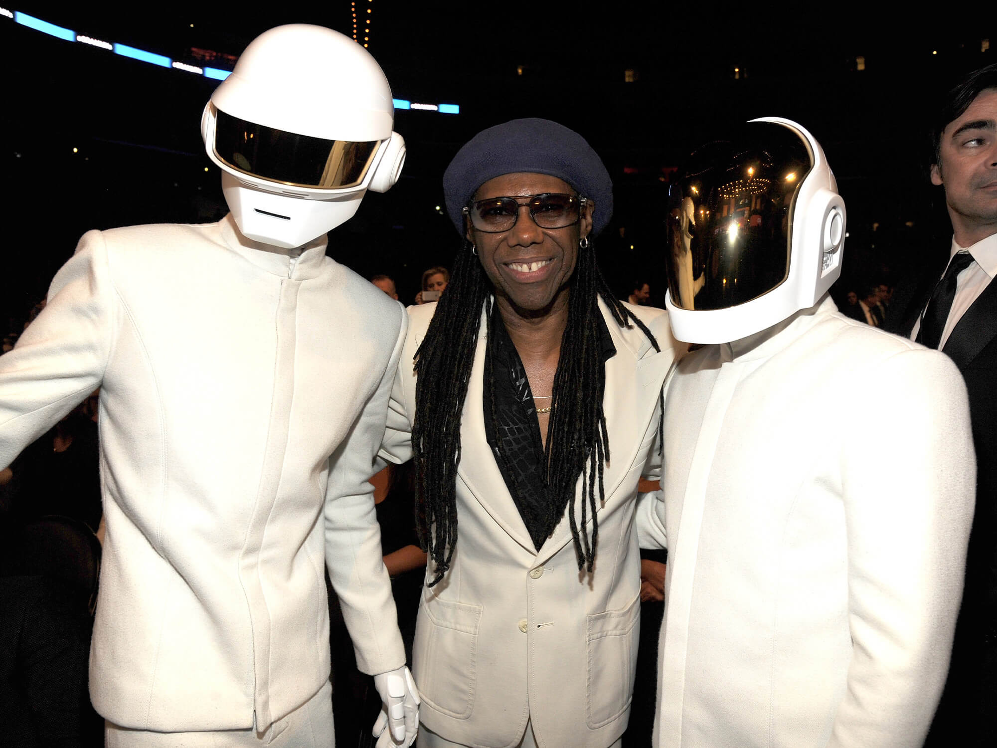 Nile Rogers stood in-between Daft Punk. All three are wearing white suits. Daft Punk are wearing their famous futuristic helmets.