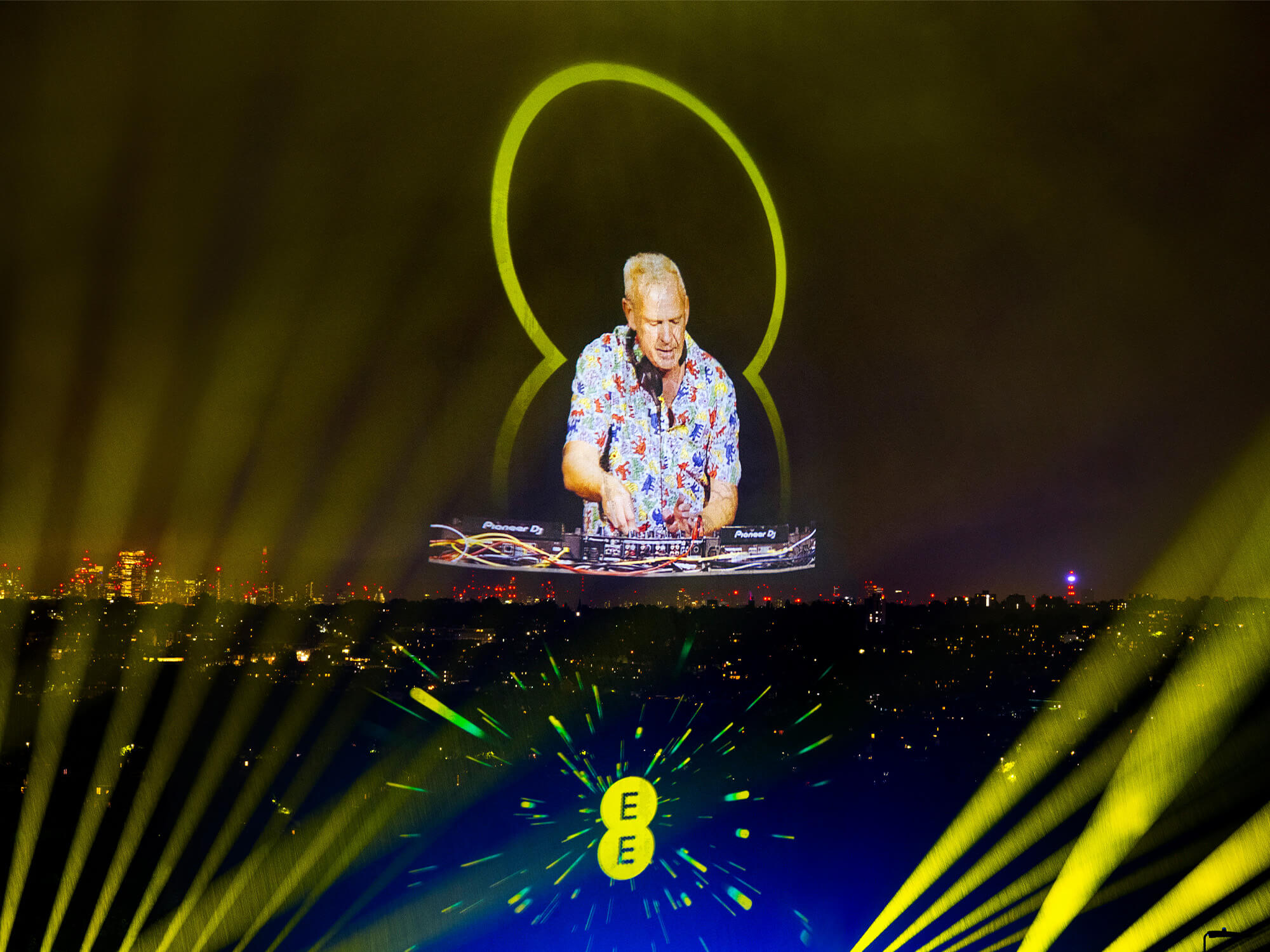 Fatboy Slim's hologram projected into the sky among yellow lights. He is stood behind decks, looking down and wearing headphones around his neck.