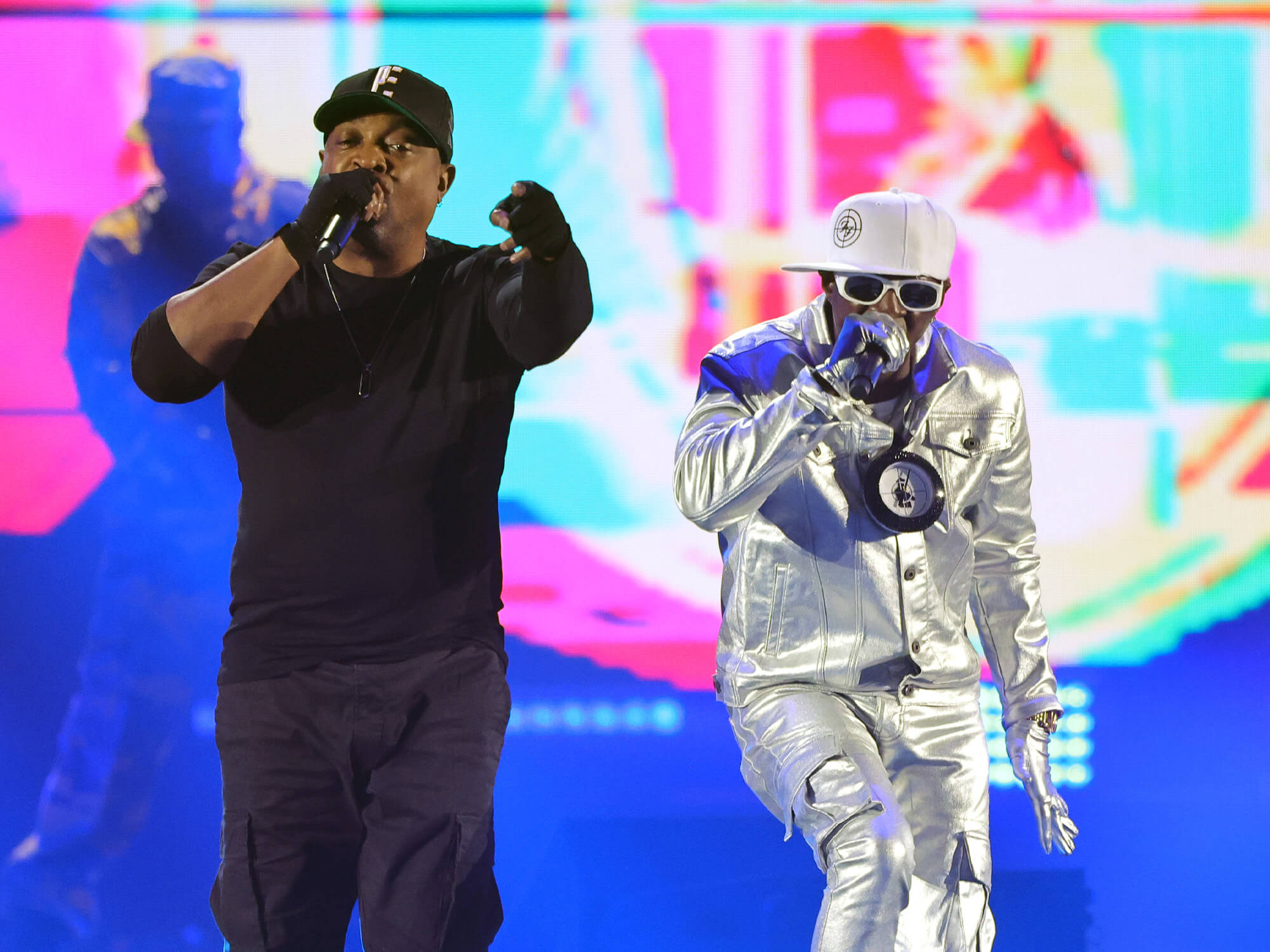 Chuck D and Flavour Flav on stage in 2023. Chuck D wears an all black outfit, whilst Flavour Flav is dressed in silver. They both hold microphones.