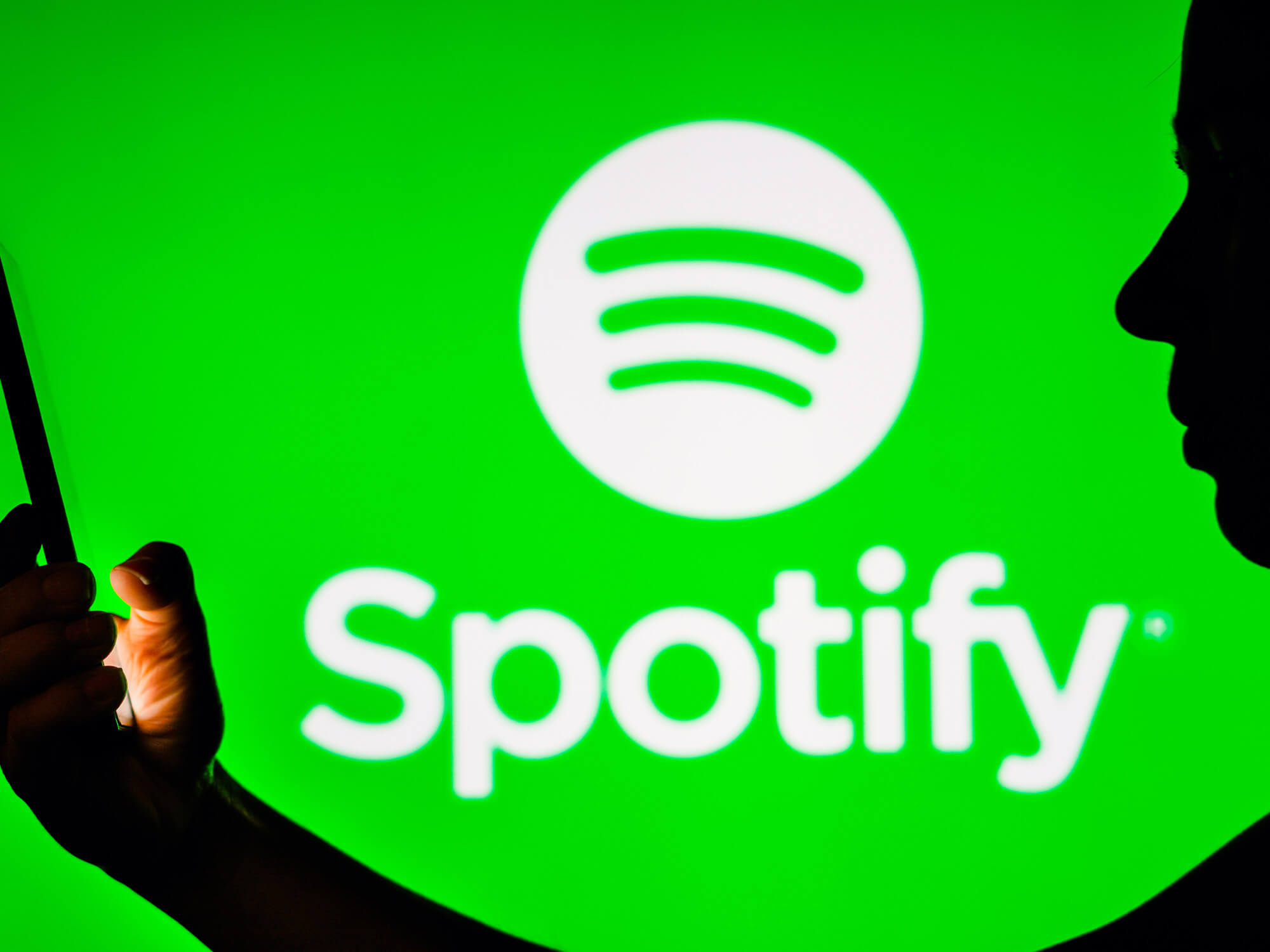 Spotify logo on green background, with silhouette of a woman holding a phone standing in front of it