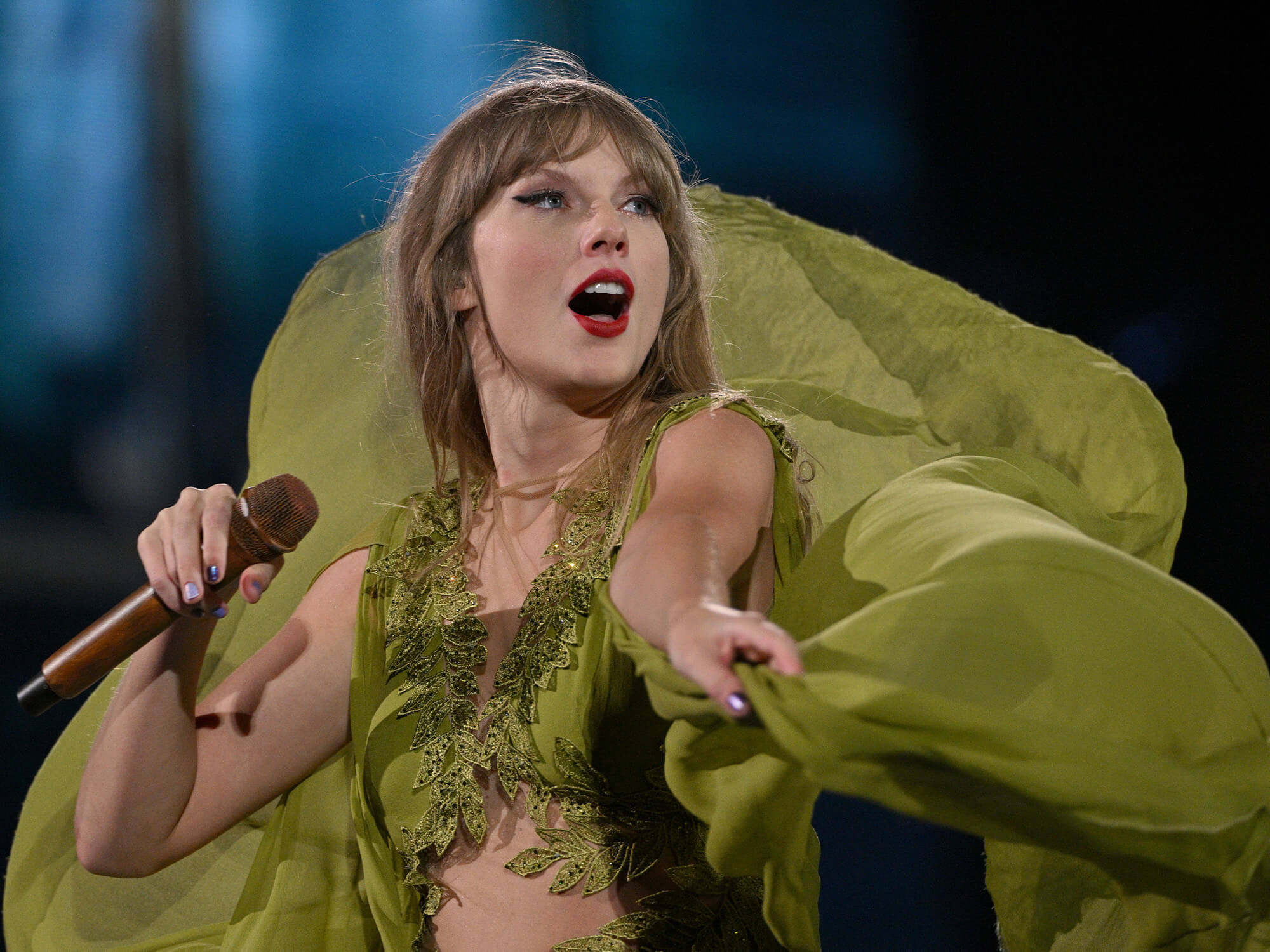 Taylor Swift on stage. She is holding a brown microphone in one hand, and holds the trail of her long green dress in the other. She is looking out into the audience.