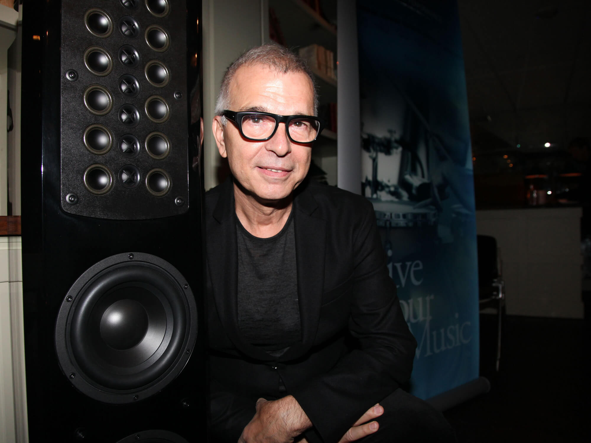 Tony Visconti sitting in front of a large speaker. He wears black-framed glasses and a black blazer jacket