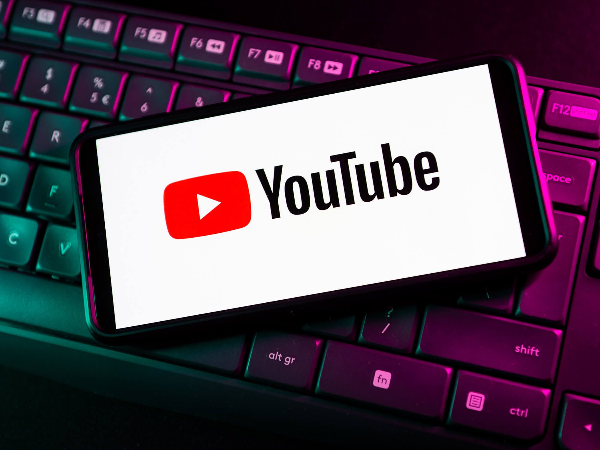 YouTube logo on a phone screen, which rests on top of a black keyboard. There is pink lighting over the keyboard.