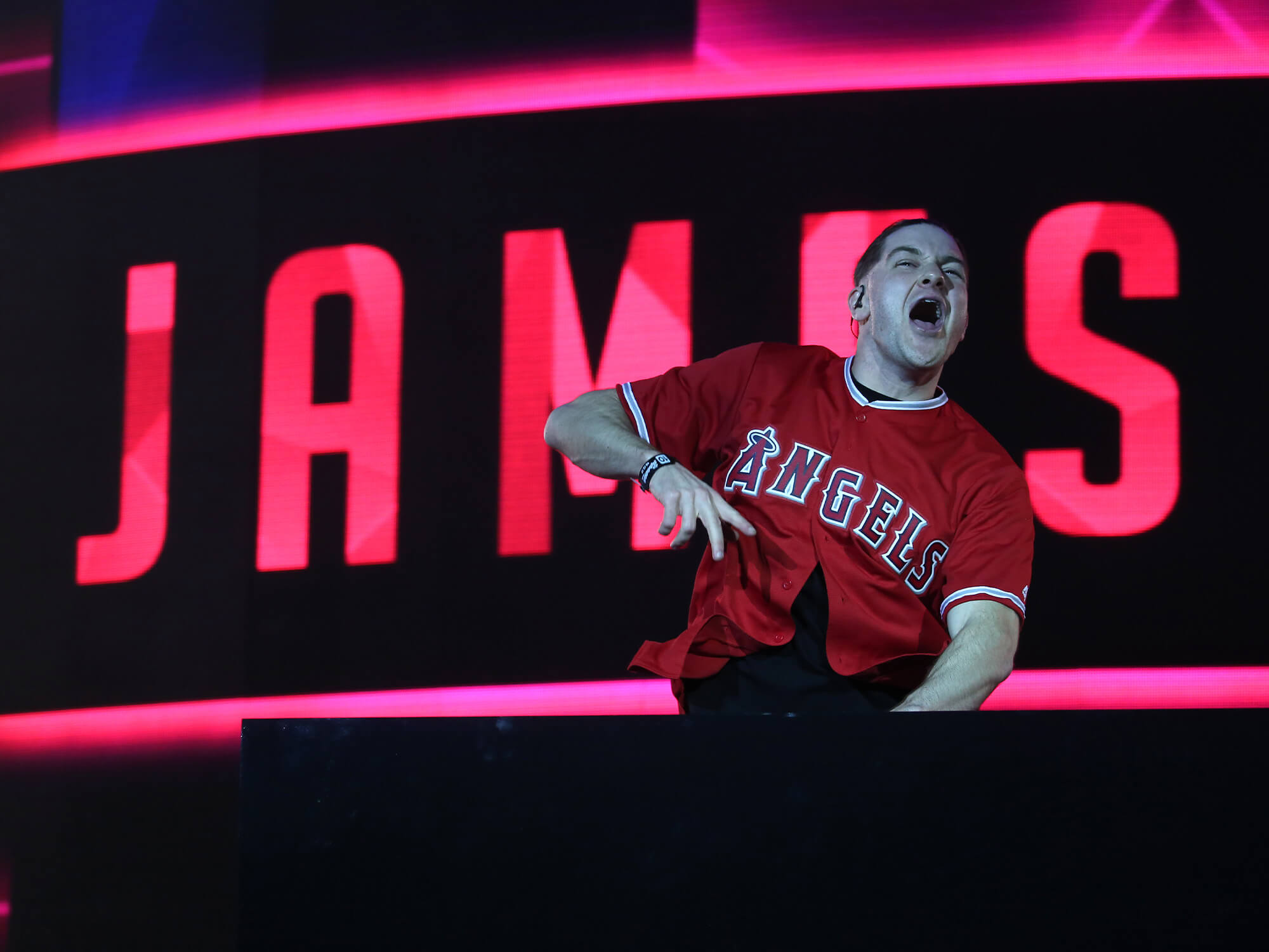James Hype on stage. He is behind the decks, is jumping in the air and is shouting out to the crowd. His name is lit up in red behind him.
