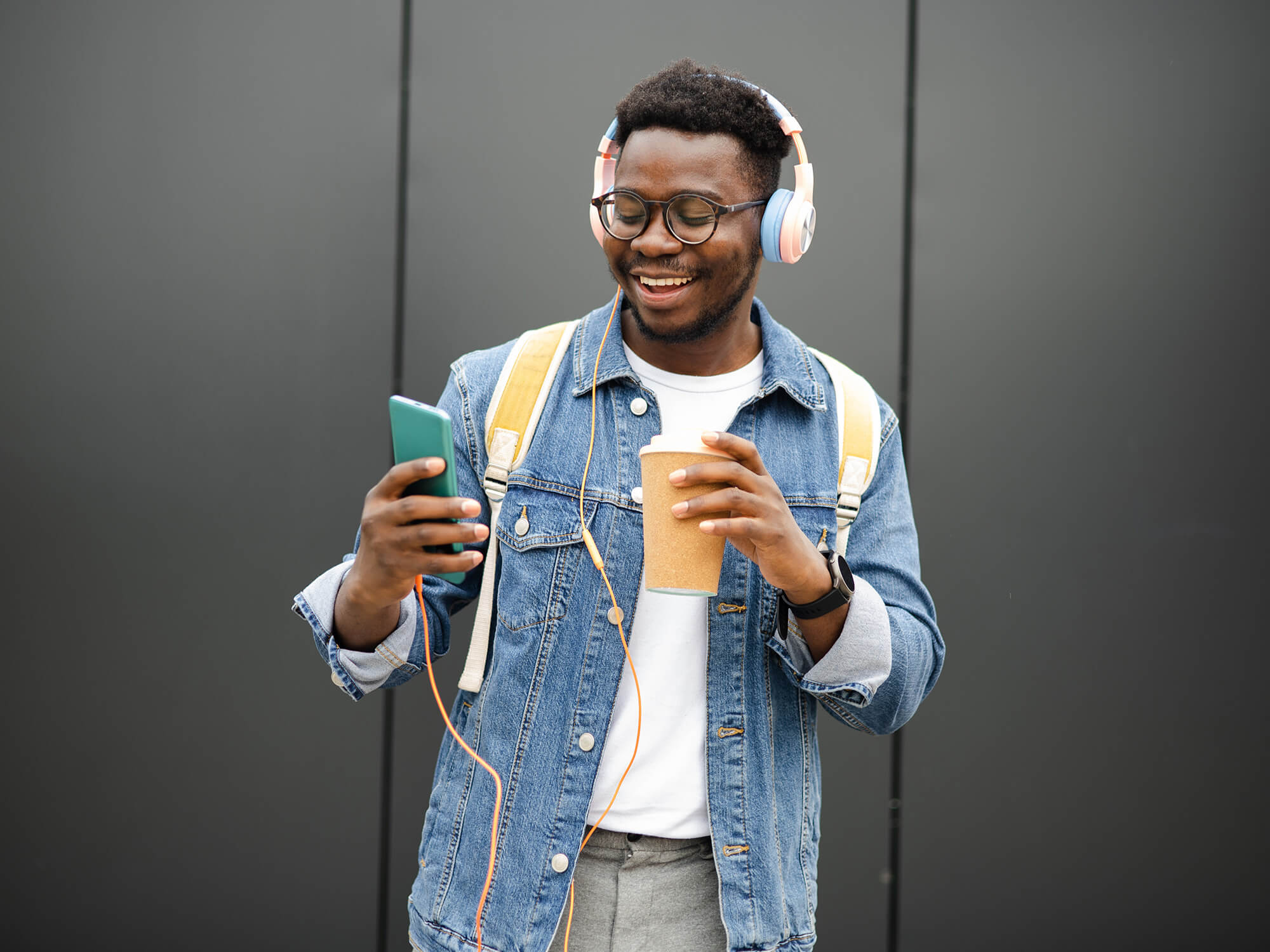 A young man listening to music through headphones and smiling. He holds his phone in one hand and drink in the other.