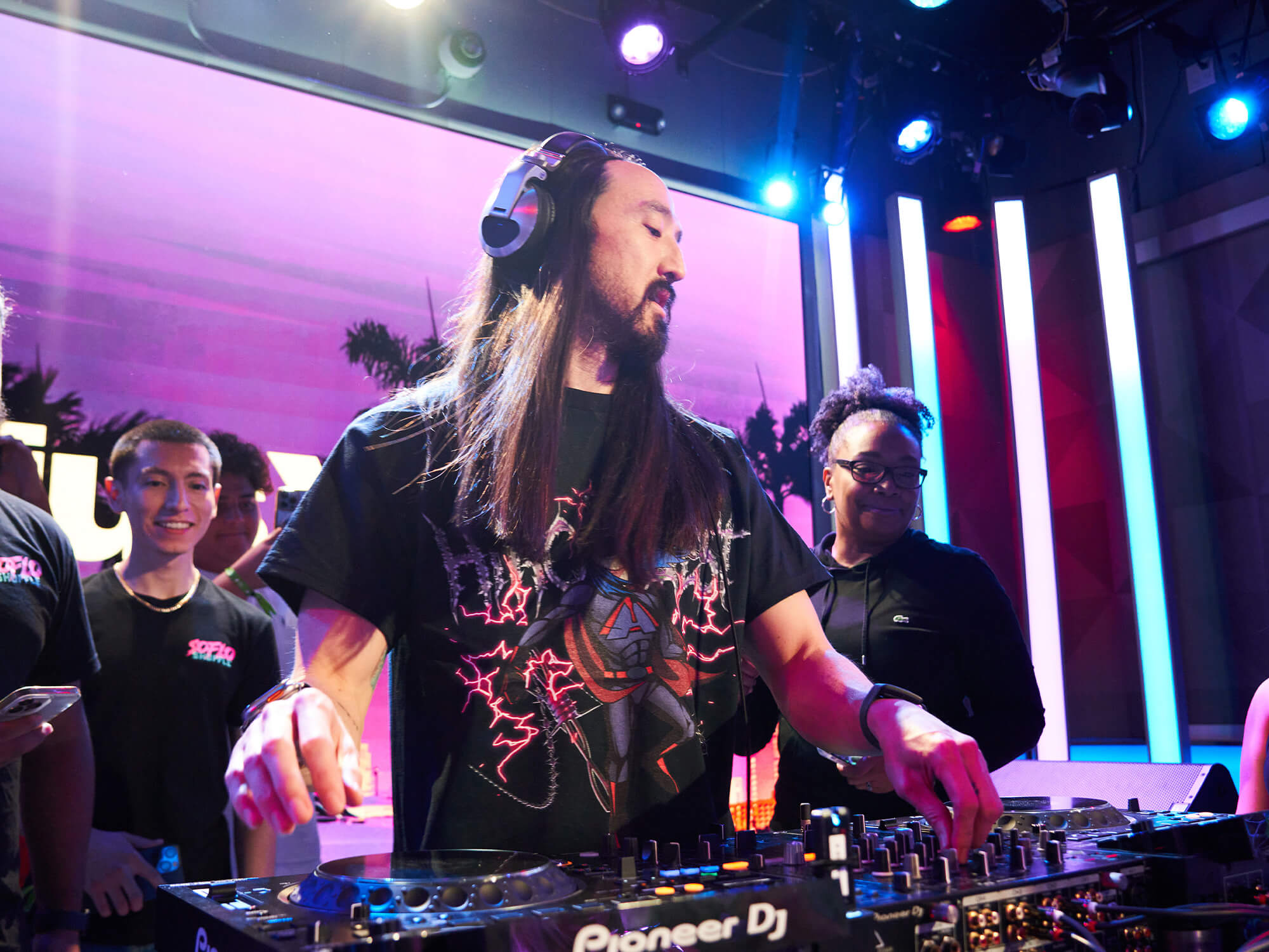 Steve Aoki behind the decks. People stand behind him and the room is lit up with purple lighting. He has long dark hair and a beard, and has headphones on.