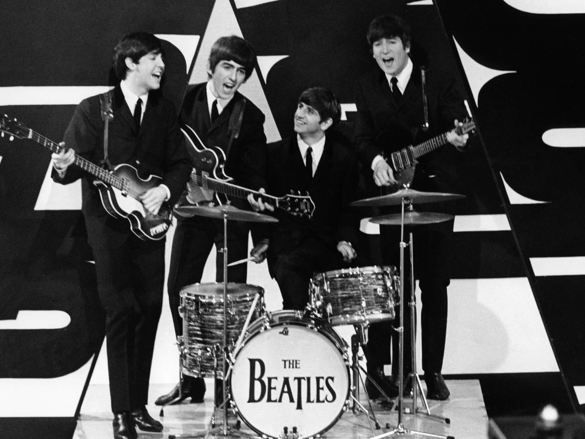 The Beatles clustered together in front of a black and white background. Paul, George and John hold their guitars and Ringo sits at his drum kit.