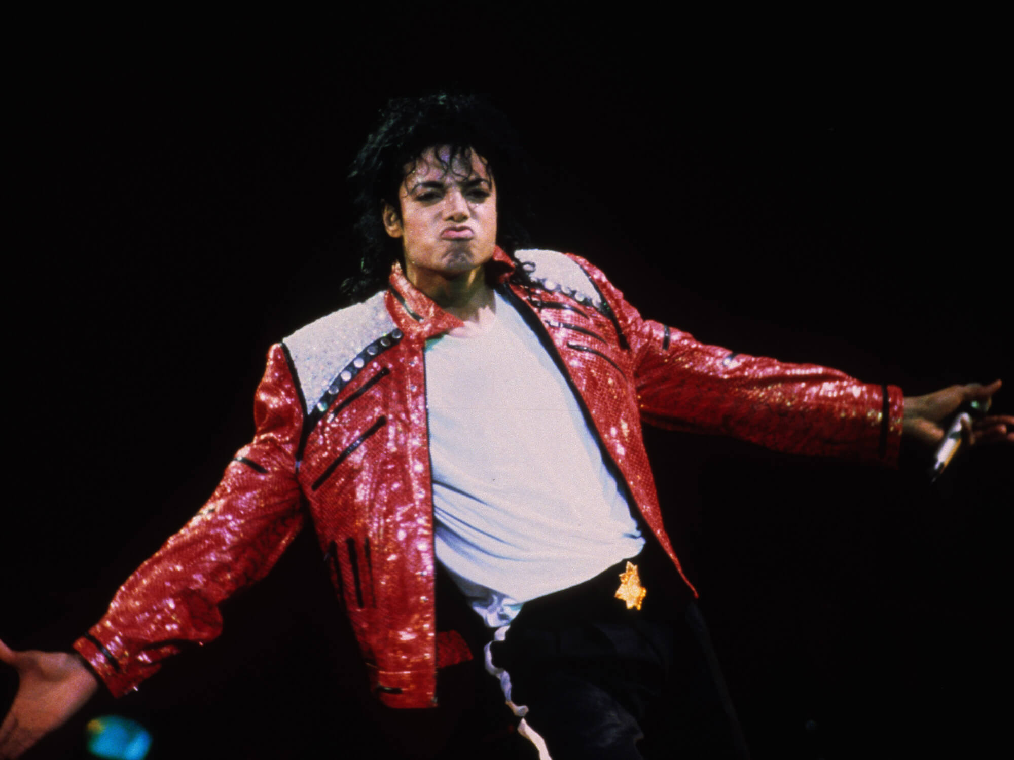 Michael Jackson on stage. He is wearing a bright leather jacket and has his arms out open to the side. He is pouting at the audience.