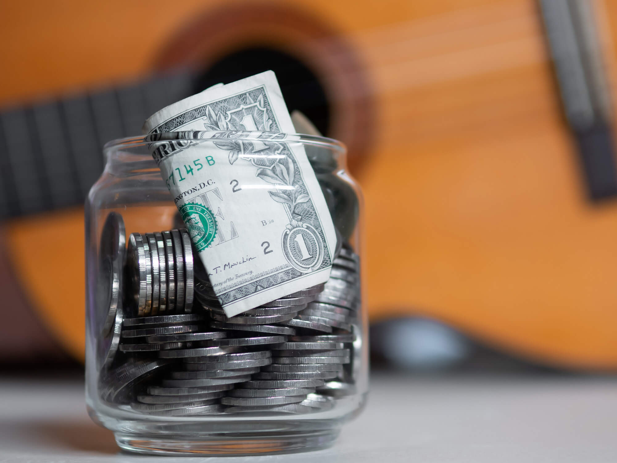 A glass jar showing silver coins an a bank note poking out of it. Blurred in the background, there is an acoustic guitar.