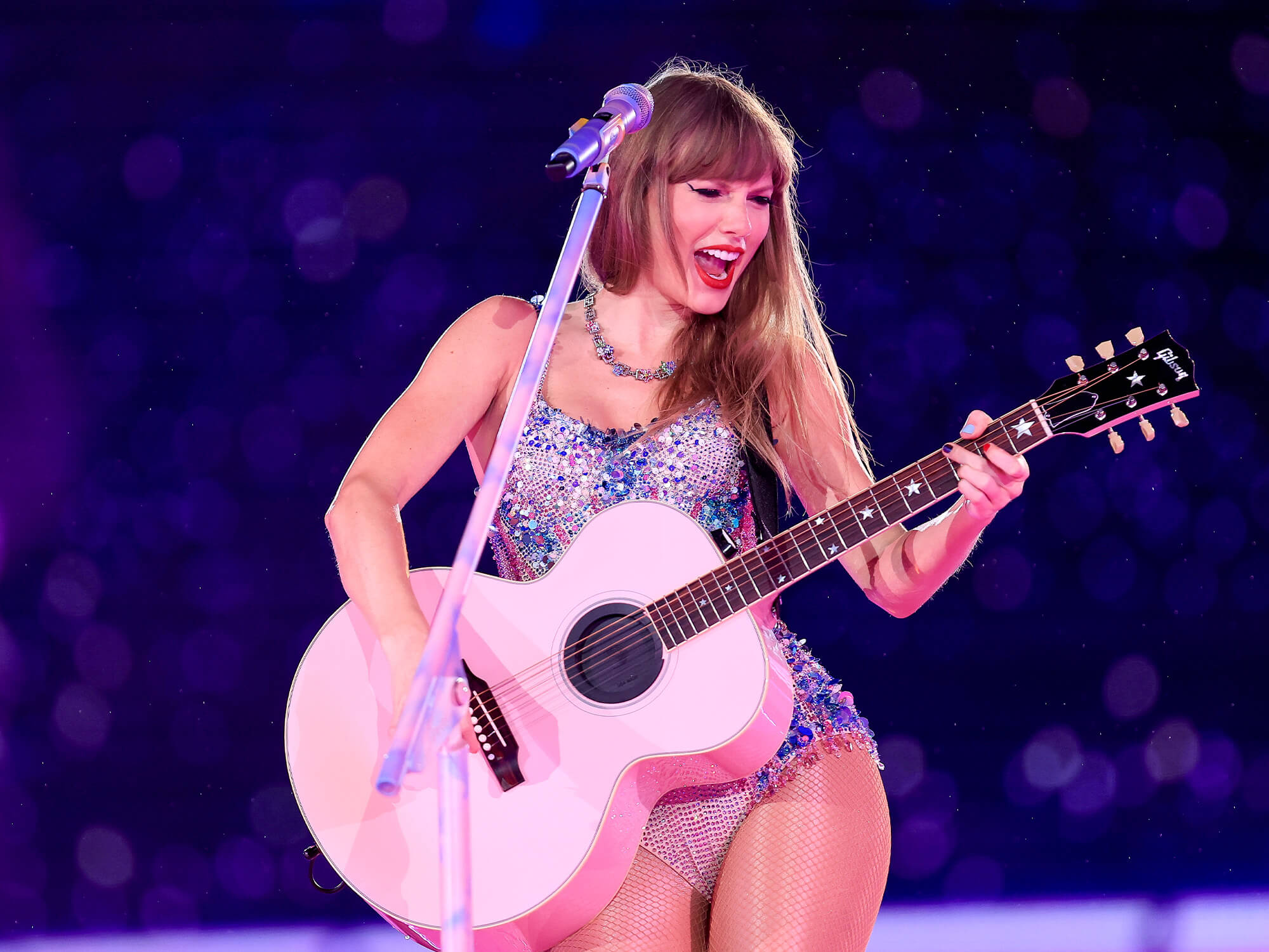 Taylor Swift playing a pink acoustic Gibson guitar on stage. She is wearing a bejewelled body suit, showing an open-mouth smile, and has long blonde hair and red lipstick.