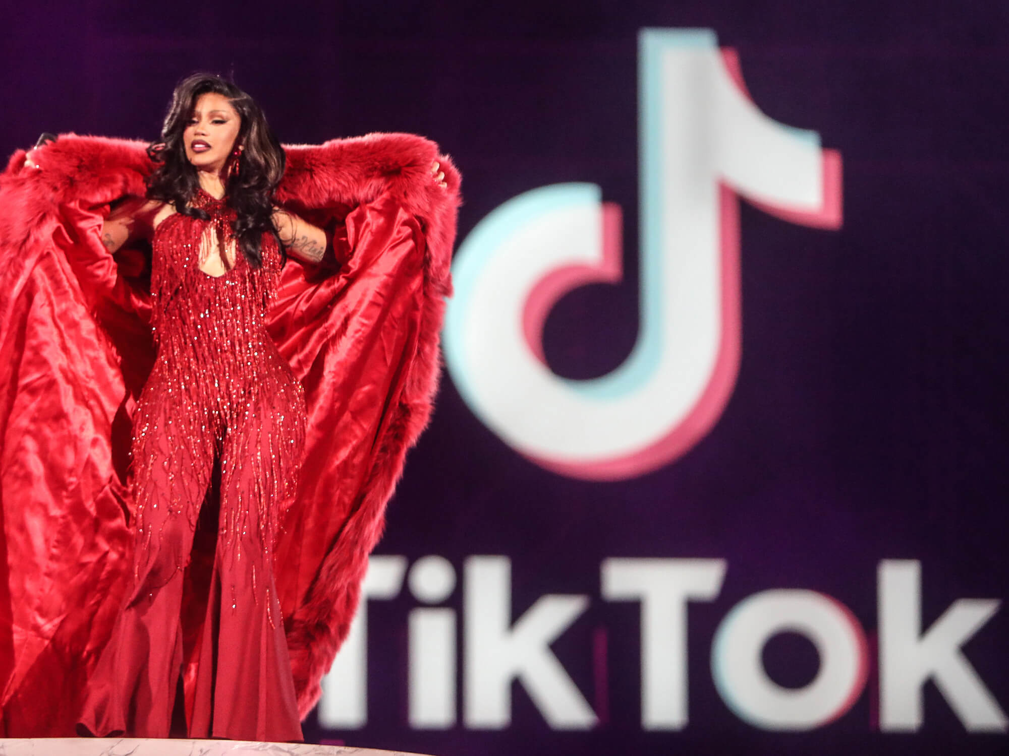 Cardi B on stage at TikTok In The Mix. She wears a vibrant red outfit and stands in front of the TikTok logo.