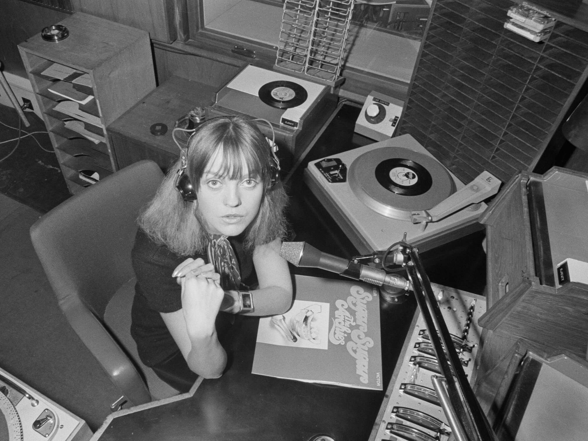 A black and white photo of Annie Nightingale taken in 1970. She is sitting in a radio studio with a set of headphones on. A mic is positioned on an metal arm in front of her.