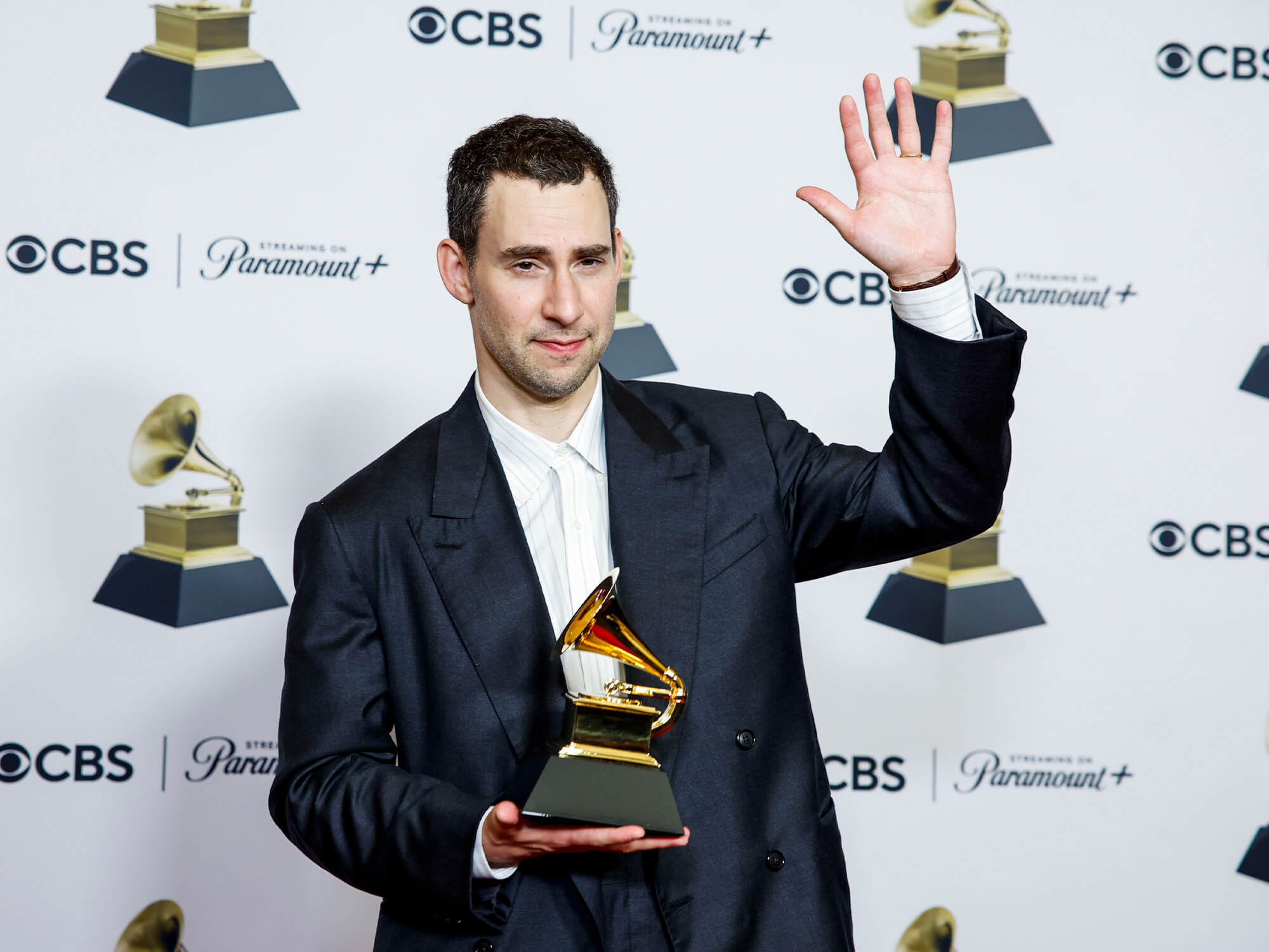 Jack Antonoff holding his Grammy award. It is a gold gramophone shape. He has one hand in the air waving.