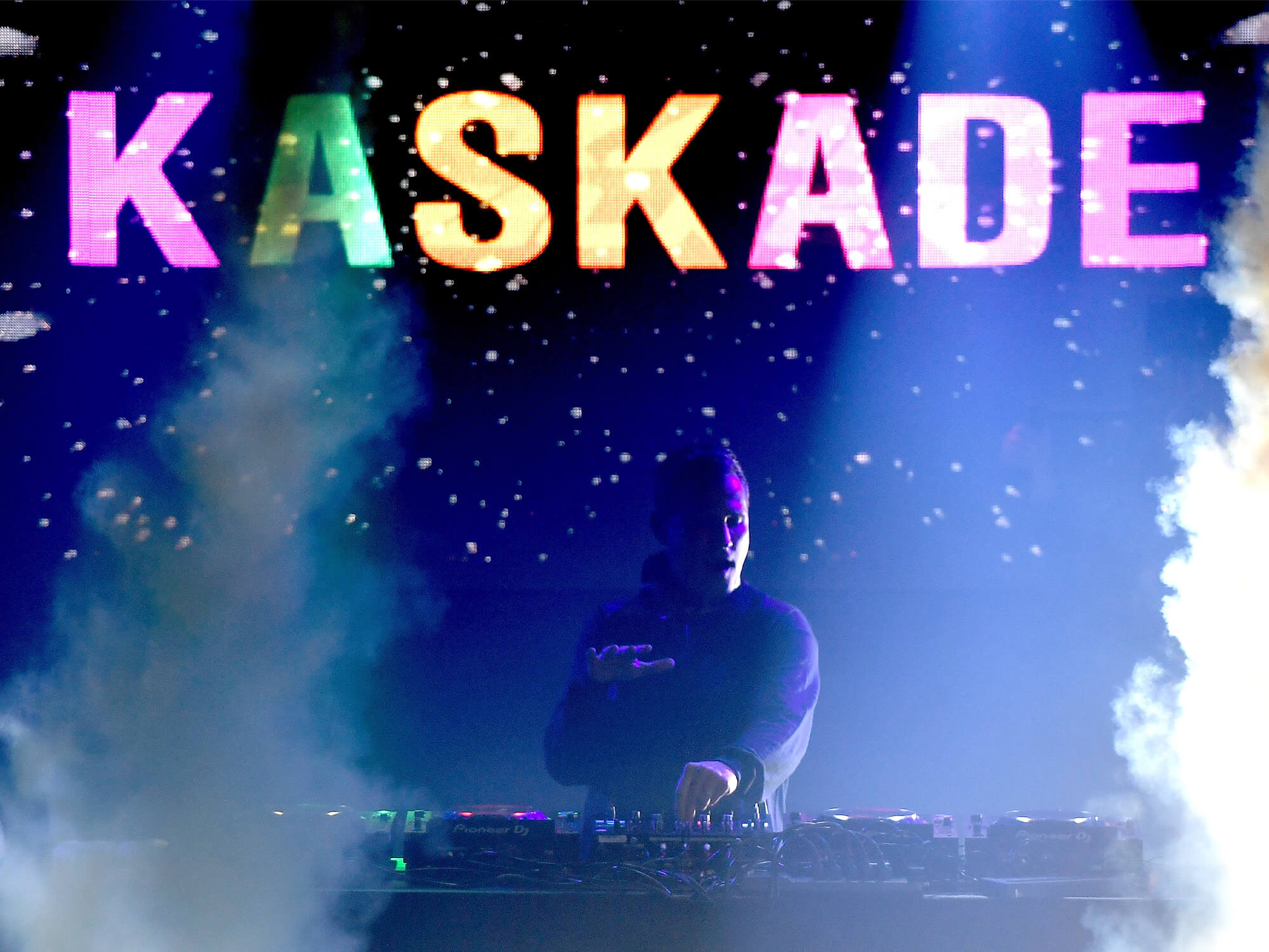 Kaskade on stage. His name is lit up in multicoloured letters, and he has on hand on a control knob on his deck.