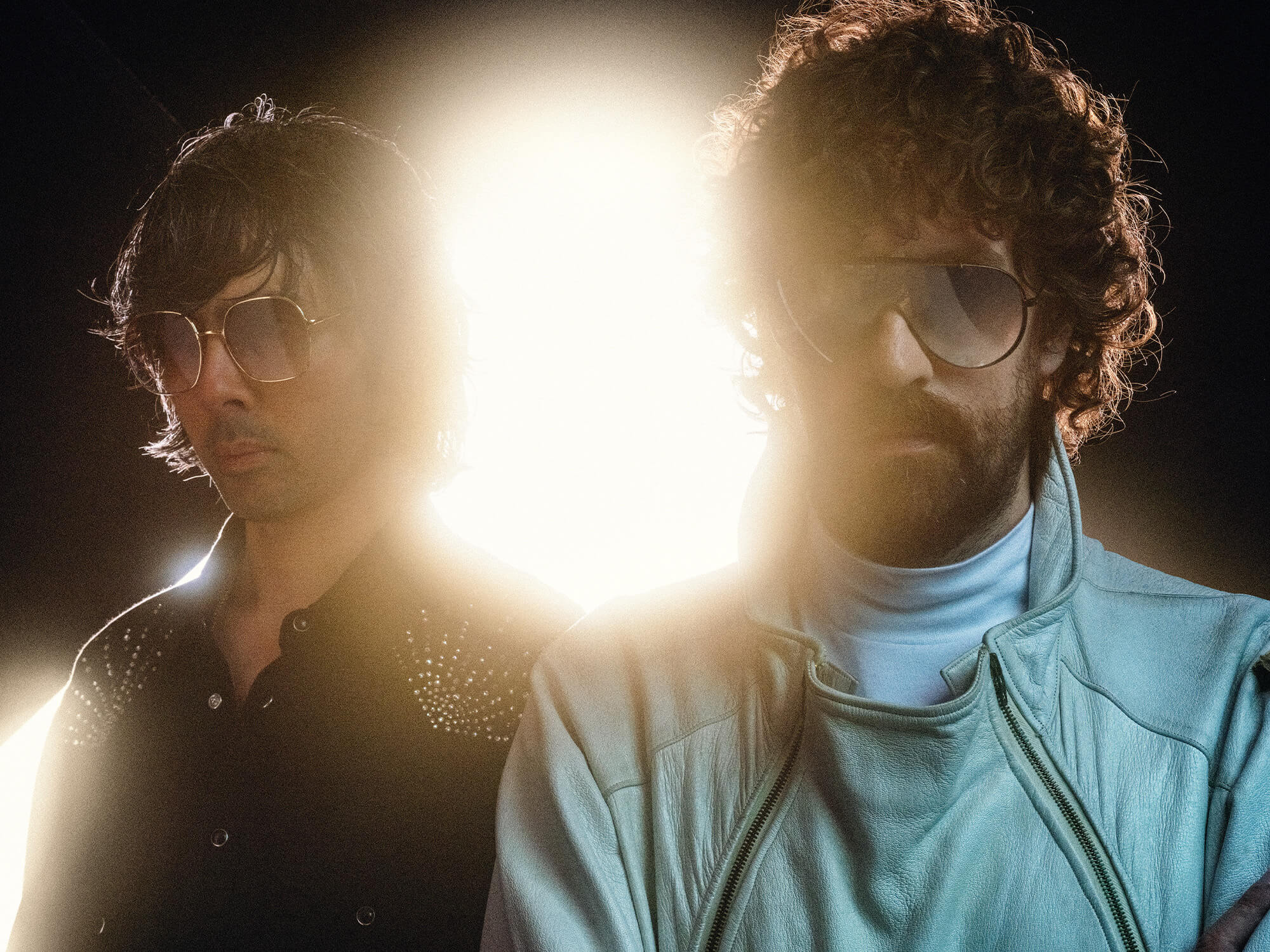 Justice standing in front of a bright white light. They are both wearing sunglasses and are looking directly at the camera.