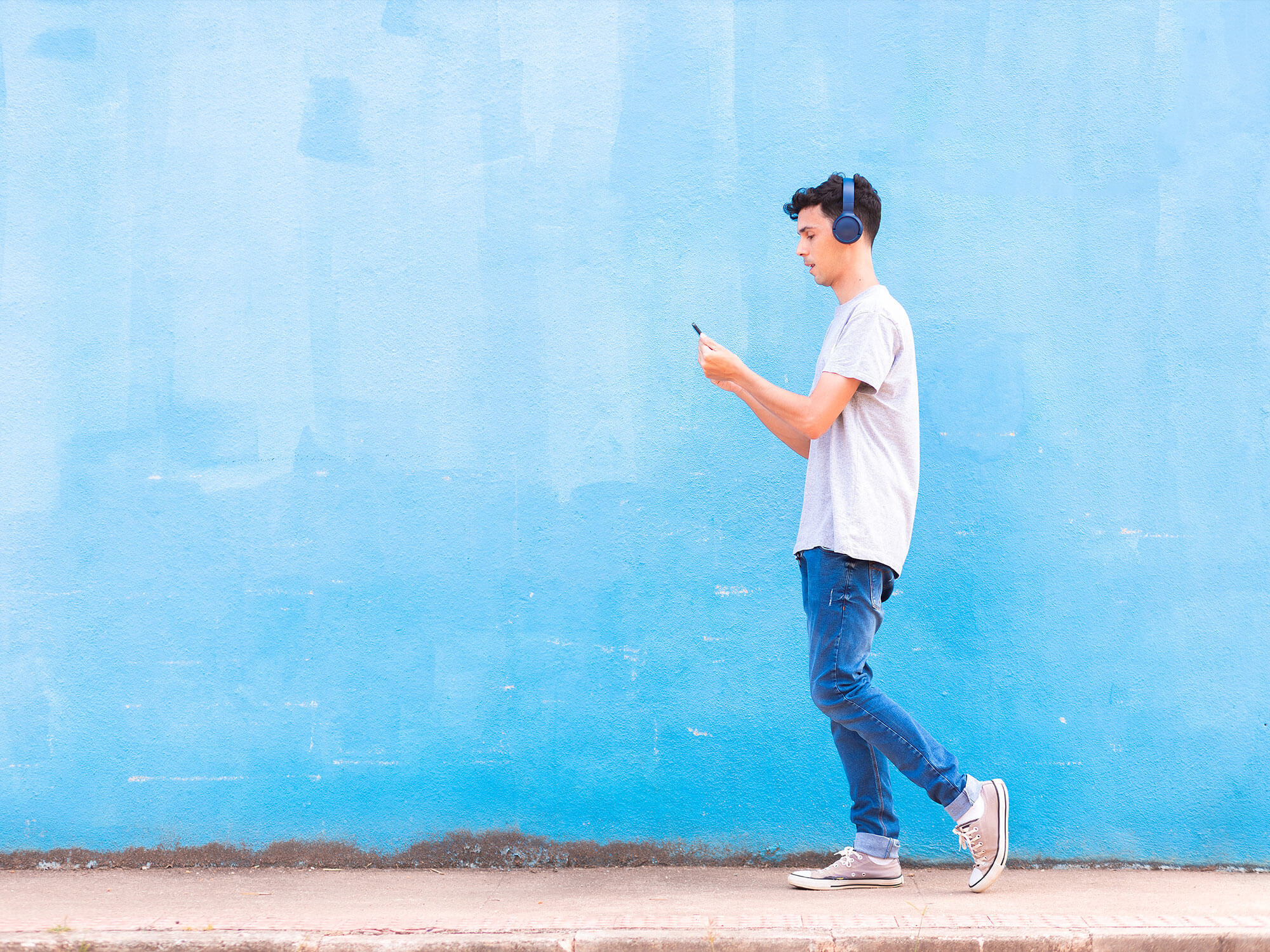 Young man wearing wireless headphones and listening to streaming music via app on his smartphone, while walking on the street sidewalk.