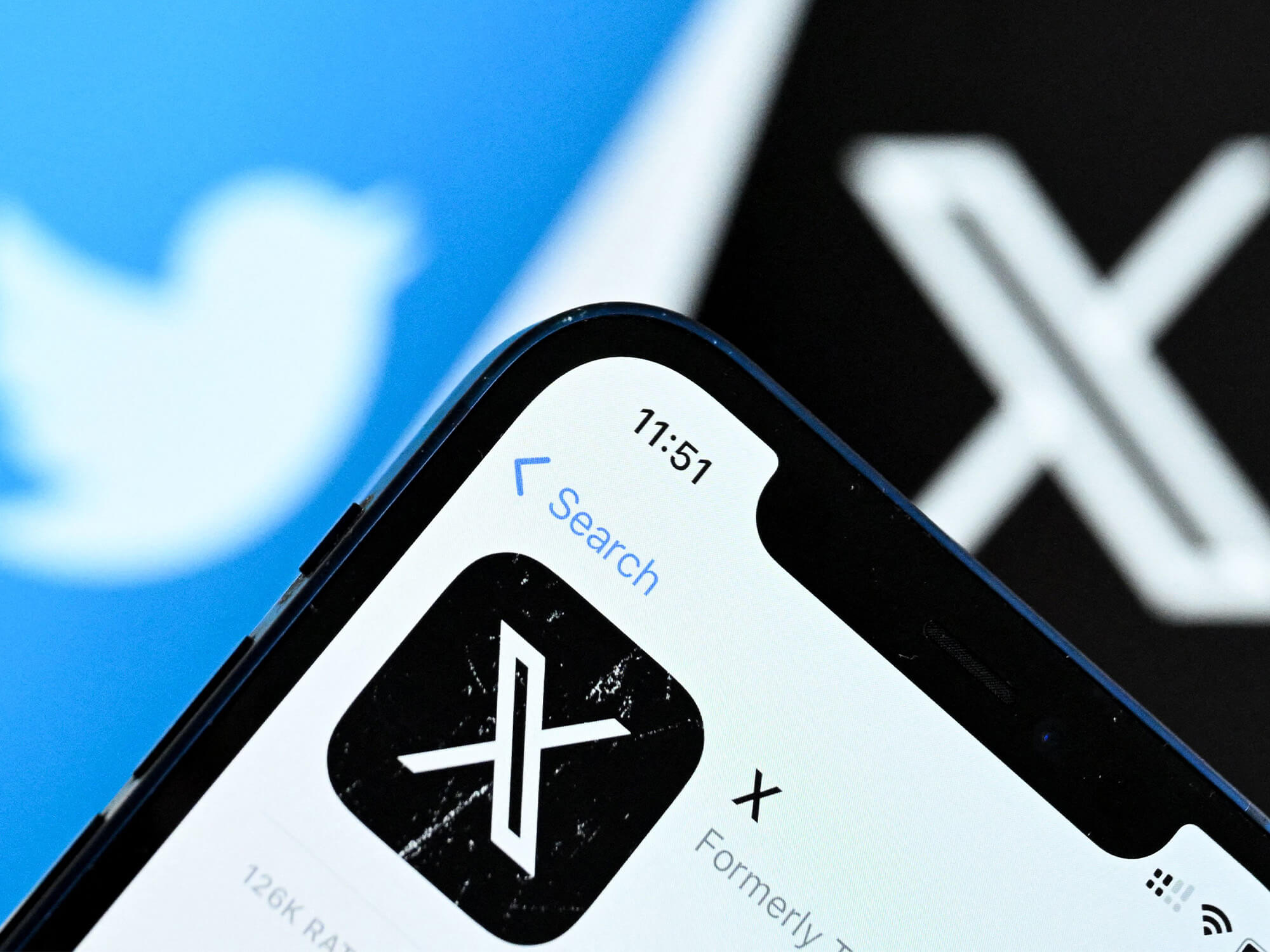 A phone showing ther X logo. Both the Twitter / X logos are blurred in the background.