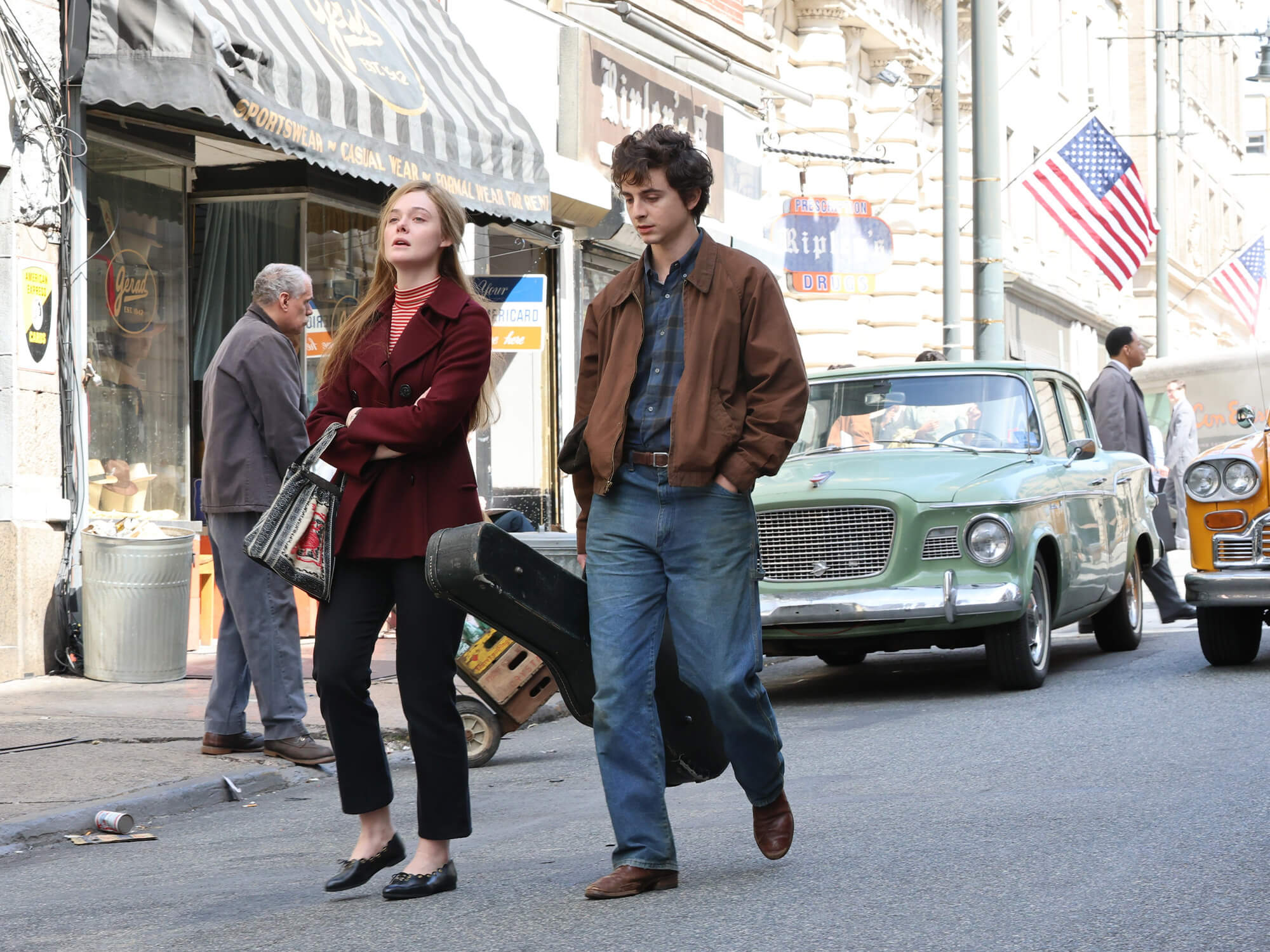 Timothee Chalamet on set of the new Bob Dylan biopic. He is captured wearing a brown jacket and holding a guitar case walking through a street.