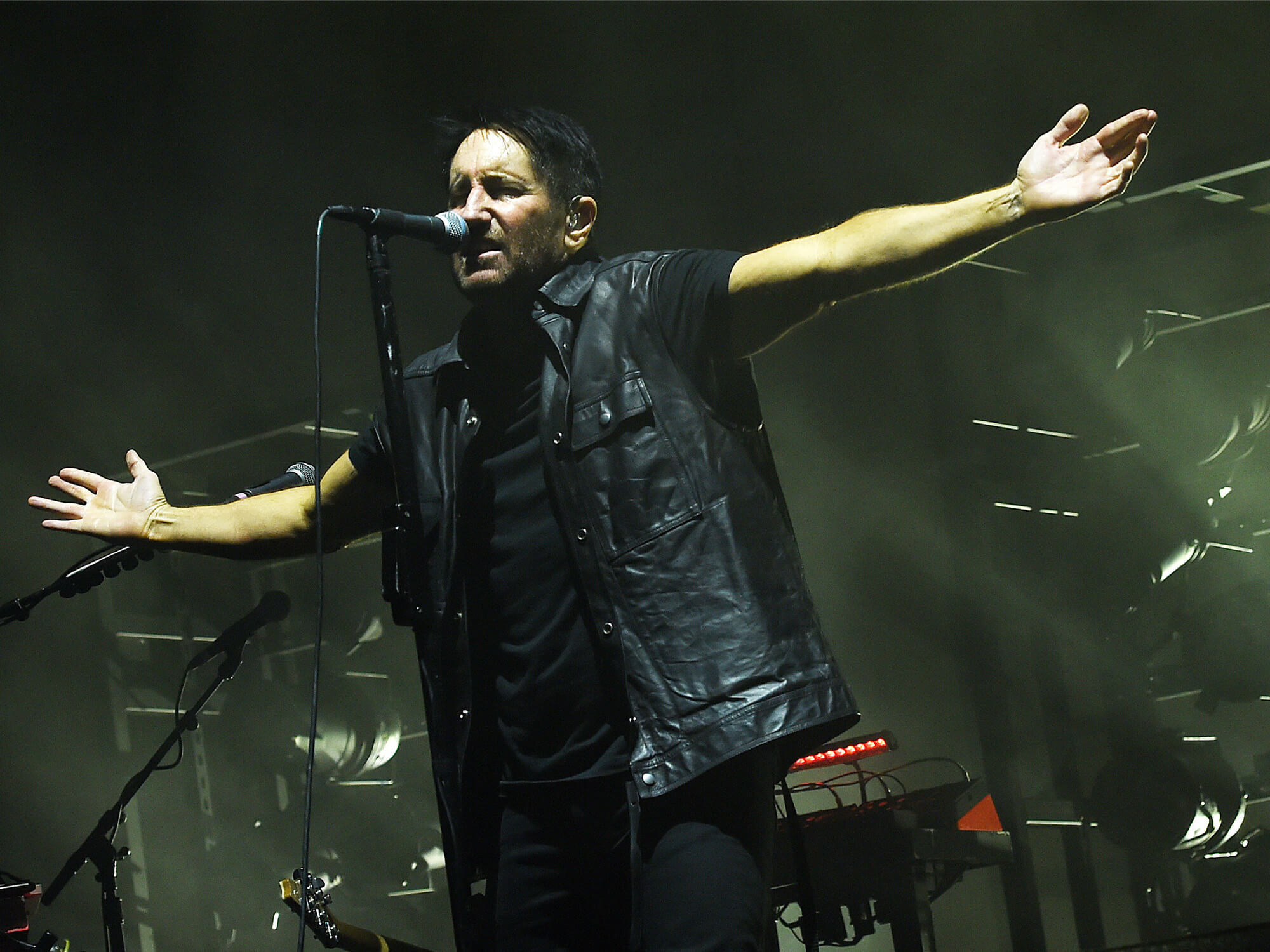 Trent Reznor on stage. He is singing into a mic on a stand and has both arms out to the side.